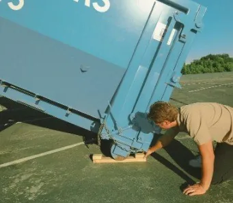 A Blue Bin driver sliding a protection board under a dumpster rail