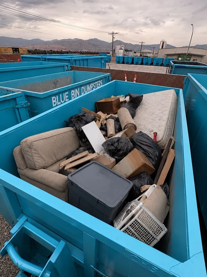 A Blue Bin dumpster loaded with a couch, a mattress, boxes and bags